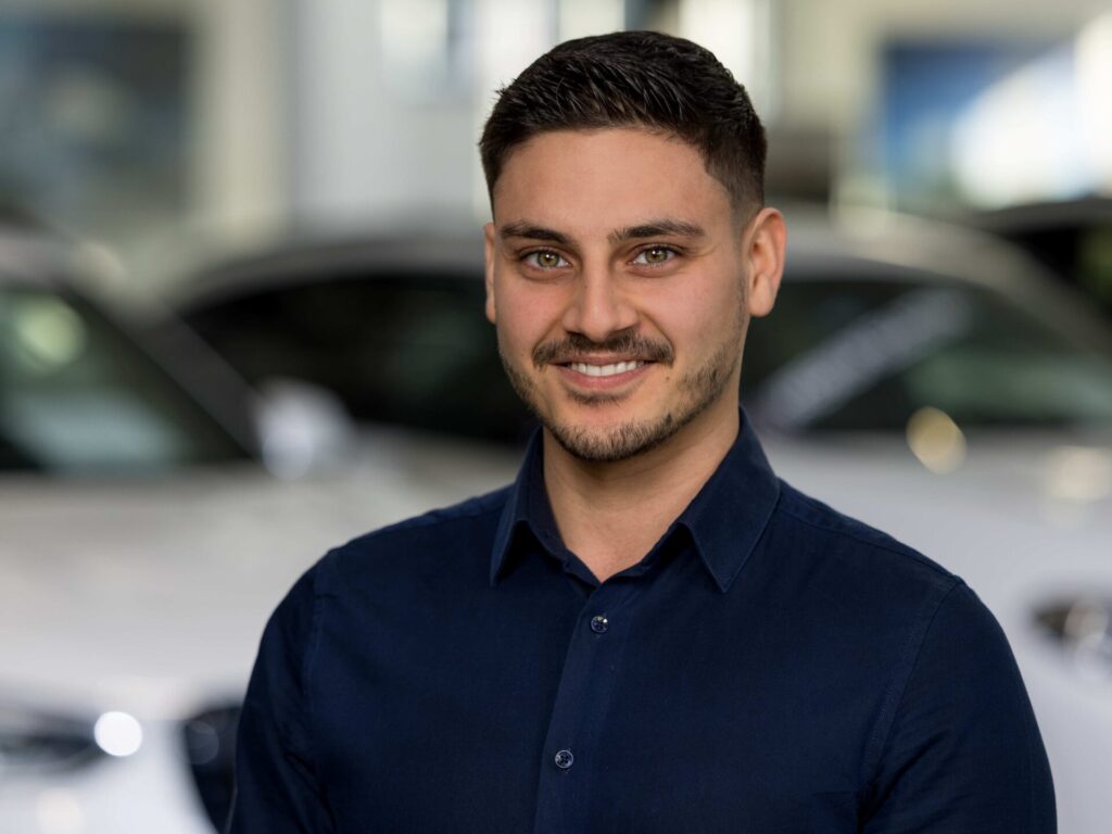 Ein Mann mit kurzen dunklen Haaren und gestutztem Bart, der ein marineblaues Button-up-Hemd trägt, lächelt in die Kamera. Im unscharfen Hintergrund sind weiße Autos in einem Ausstellungsraum zu sehen, die auf Ihren Ansprechpartner für alle Fragen rund ums Auto hinweisen. Rider's Point der Kaltenbach Gruppe. Dein Partner für BMW Motorrad und Honda.