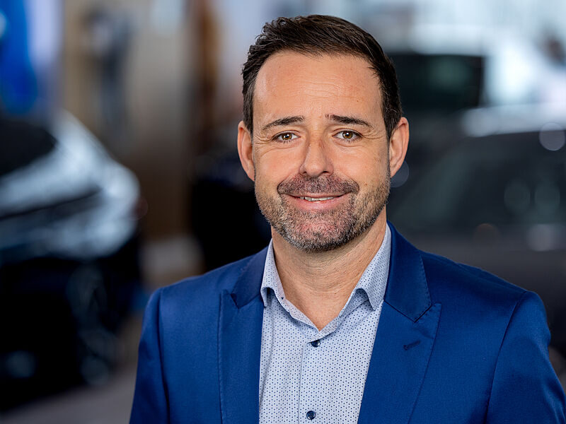 Ein Mann mit kurzen dunklen Haaren und einem Bart, der eine blaue Anzugsjacke und ein hellblaues Hemd trägt, lächelt in die Kamera - ein idealer Ansprechpartner für Alle in einem Innenraum mit unscharfem Hintergrund. Rider's Point der Kaltenbach Gruppe. Dein Partner für BMW Motorrad und Honda.