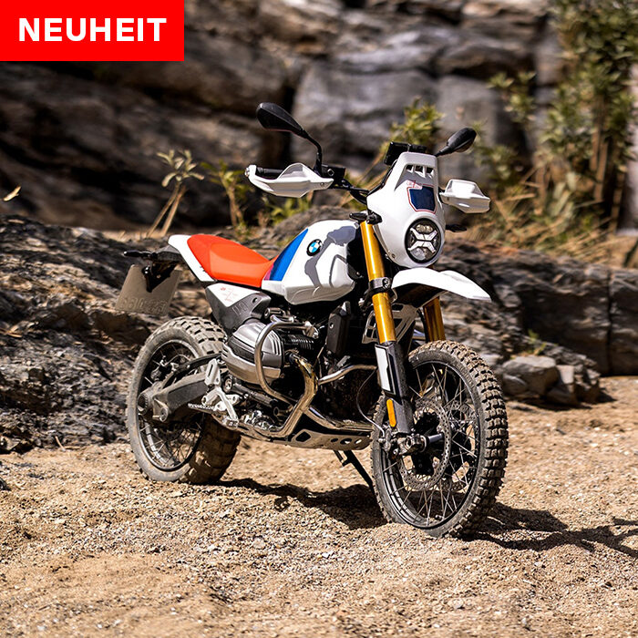 Ein BMW Adventure-Motorrad mit Geländereifen steht auf einem Feldweg in der Nähe von felsigem Gelände. Die in Weiß, Blau und Rot gehaltene NEUHEIT ist bereit für die BMW Probefahrtwochen Werdohl. Rider's Point der Kaltenbach Gruppe. Dein Partner für BMW Motorrad und Honda.
