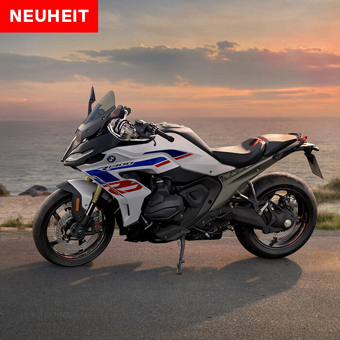 Ein modernes Sporttouring-Motorrad mit roten, blauen und schwarzen Akzenten ist bei Sonnenuntergang an einer Küstenstraße geparkt. Der Himmel ist bunt, im Hintergrund schimmert das Meer, und ein rotes NEUHEIT-Schild weist auf die BMW Probefahrtwochen Werdohl hin. Rider's Point der Kaltenbach Gruppe. Dein Partner für BMW Motorrad und Honda.