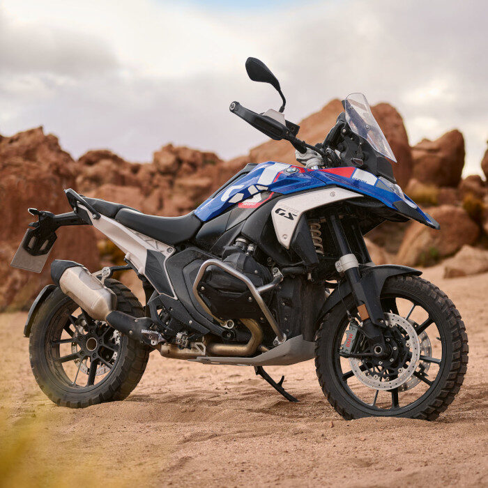 Ein blau-weiß-rotes BMW Adventure-Motorrad parkt während der BMW Probefahrtwochen Werdohl auf sandigem Gelände mit Felsformationen im Hintergrund unter bewölktem Himmel. Rider's Point der Kaltenbach Gruppe. Dein Partner für BMW Motorrad und Honda.