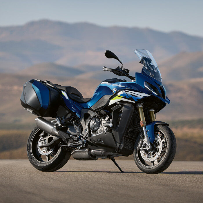 Ein blaues Sporttouring-Motorrad mit Hartschalenkoffern ist während der BMW Probefahrtwochen Werdohl auf einer Straße geparkt, mit verschwommenen Bergen und einem klaren Himmel im Hintergrund. Rider's Point der Kaltenbach Gruppe. Dein Partner für BMW Motorrad und Honda.