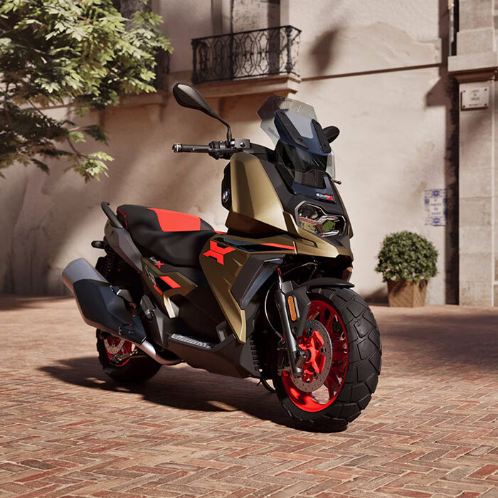 Ein modernes, sportliches Motorrad mit gold-schwarzer Karosserie, roten Akzenten und dicken Reifen parkt während der BMW Probefahrtwochen Werdohl auf einer sonnenbeschienenen Backsteinstraße in der Nähe eines Gebäudes mit Balkonen und Topfpflanzen. Rider's Point der Kaltenbach Gruppe. Dein Partner für BMW Motorrad und Honda.