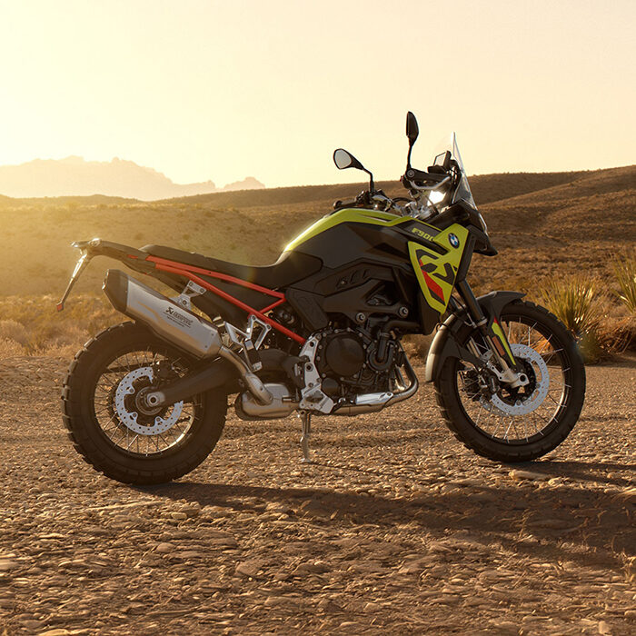 Ein gelb-schwarzes Adventure-Motorrad, geparkt auf einem felsigen Wüstenpfad bei Sonnenuntergang, mit karger Vegetation und fernen Bergen im Hintergrund - perfekt für eine Erkundungstour vor den BMW Probefahrtwochen Werdohl. Rider's Point der Kaltenbach Gruppe. Dein Partner für BMW Motorrad und Honda.