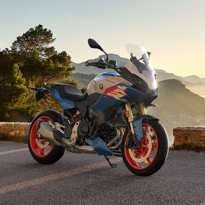Ein modernes blau-weiß-rotes BMW Motorrad, geparkt auf einer malerischen Bergstraße bei Sonnenuntergang, umgeben von Bäumen und fernen Hügeln unter einem klaren Himmel - perfekt für Ihr BMW Probefahrtwochen Werdohl Erlebnis. Rider's Point der Kaltenbach Gruppe. Dein Partner für BMW Motorrad und Honda.