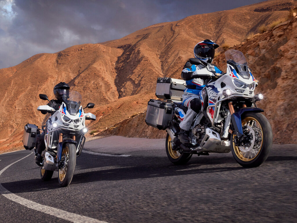 Zwei Motorradfahrer fahren mit Abenteuermotorrädern und Gepäckboxen auf einer kurvenreichen Bergstraße und präsentieren ihren Fahrstil inmitten von felsigen, wüstenartigen Hügeln unter einem wolkenverhangenen Himmel - eine perfekte Szene für das Saisonfinale 2025. Rider's Point der Kaltenbach Gruppe. Dein Partner für BMW Motorrad und Honda.