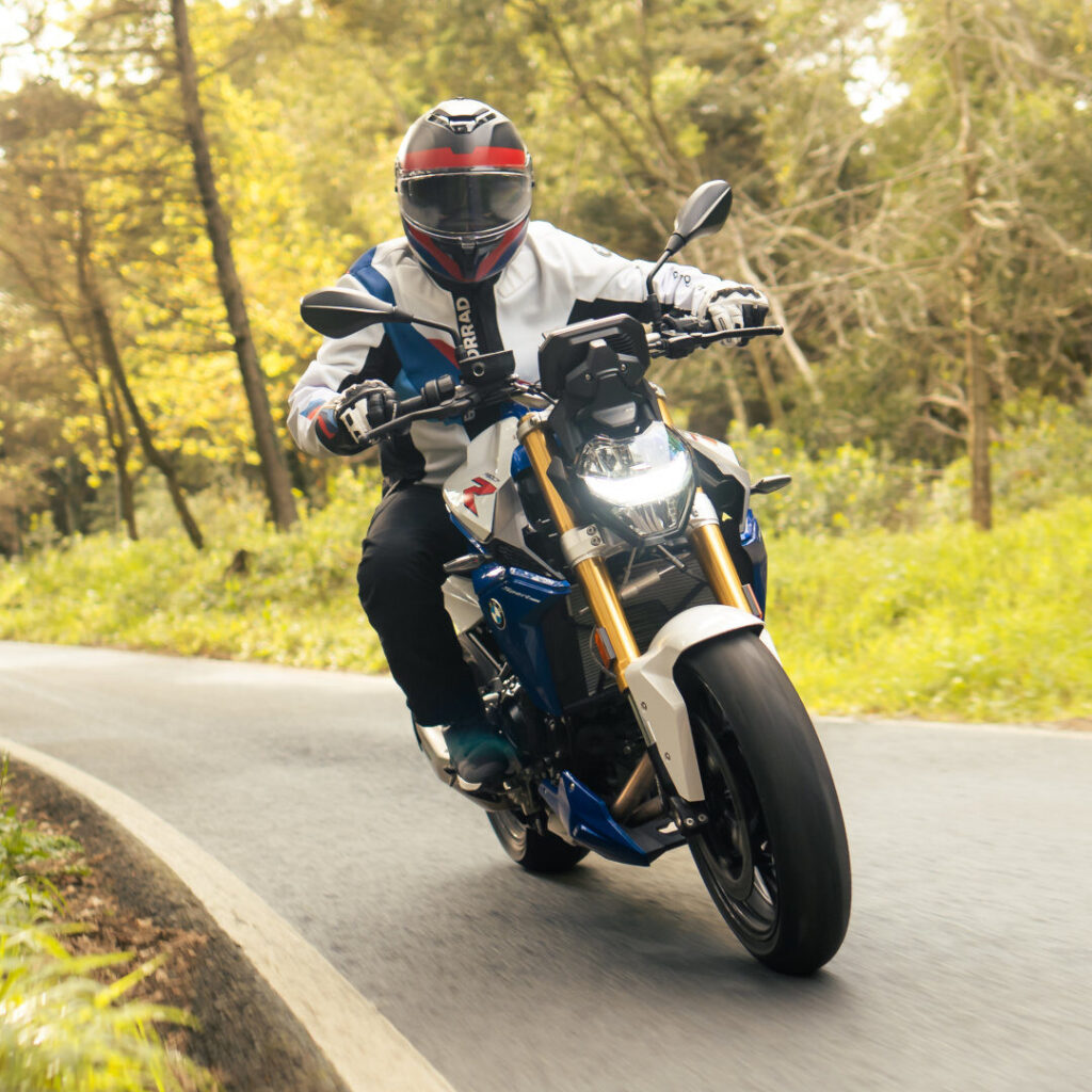 Ein Motorradfahrer mit Helm und Schutzausrüstung fährt mit seiner blau-weißen BMW F 900 R auf einer kurvenreichen Straße durch ein Waldgebiet mit üppigem Grün. Rider's Point der Kaltenbach Gruppe. Dein Partner für BMW Motorrad und Honda.