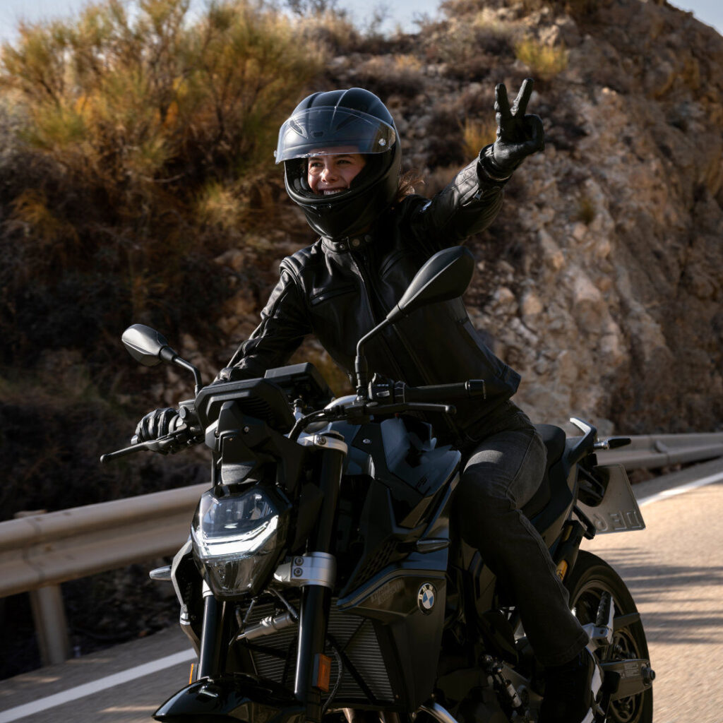 Eine Person, die einen schwarzen Helm und eine Lederjacke trägt, fährt mit einem BMW F 900 R Motorrad auf einer Straße neben einem felsigen Hügel, lächelt und macht mit der rechten Hand ein Friedenszeichen. Rider's Point der Kaltenbach Gruppe. Dein Partner für BMW Motorrad und Honda.