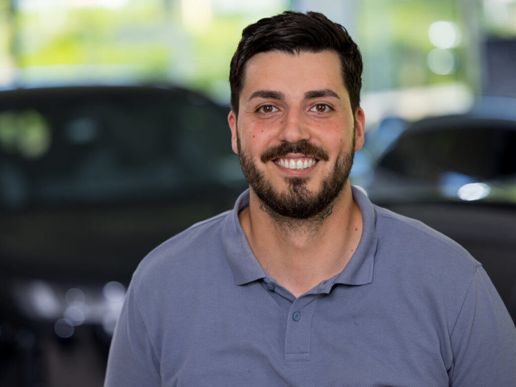 Ein Mann mit dunklen Haaren und Bart, der ein hellgraues Polohemd trägt, lächelt in die Kamera, während im Hintergrund Autos und Grünflächen verschwimmen - ideal für eine Beitragsseite oder als Bild für einen Ansprechpartner Verkauf. Rider's Point der Kaltenbach Gruppe. Dein Partner für BMW Motorrad und Honda.
