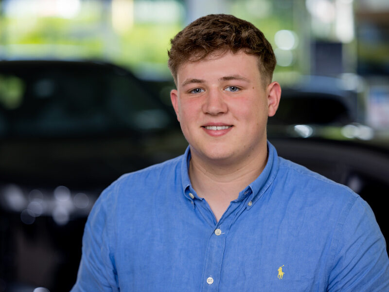 Ein junger Mann mit kurzen braunen Haaren, der ein blaues Button-up-Hemd trägt, lächelt in die Kamera. Der unscharfe Hintergrund deutet auf Autos im Innenraum hin - perfekt für einen Ansprechpartner im Teilevertrieb. Rider's Point der Kaltenbach Gruppe. Dein Partner für BMW Motorrad und Honda.