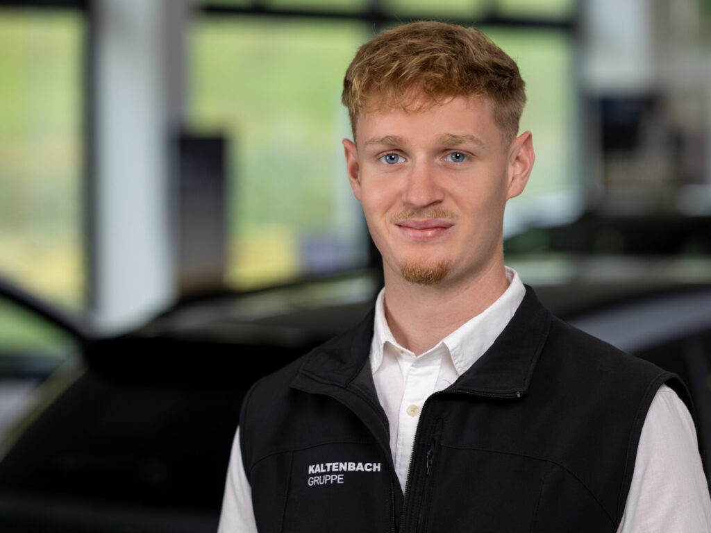 Ein junger Mann mit kurzen blonden Haaren und gestutztem Bart, der ein weißes Hemd und eine schwarze Weste mit der Aufschrift Kaltenbach Gruppe trägt, steht in einem Haus vor einem verschwommenen Hintergrund mit Autos - Ihr Ansprechpartner im Teilevertrieb. Rider's Point der Kaltenbach Gruppe. Dein Partner für BMW Motorrad und Honda.