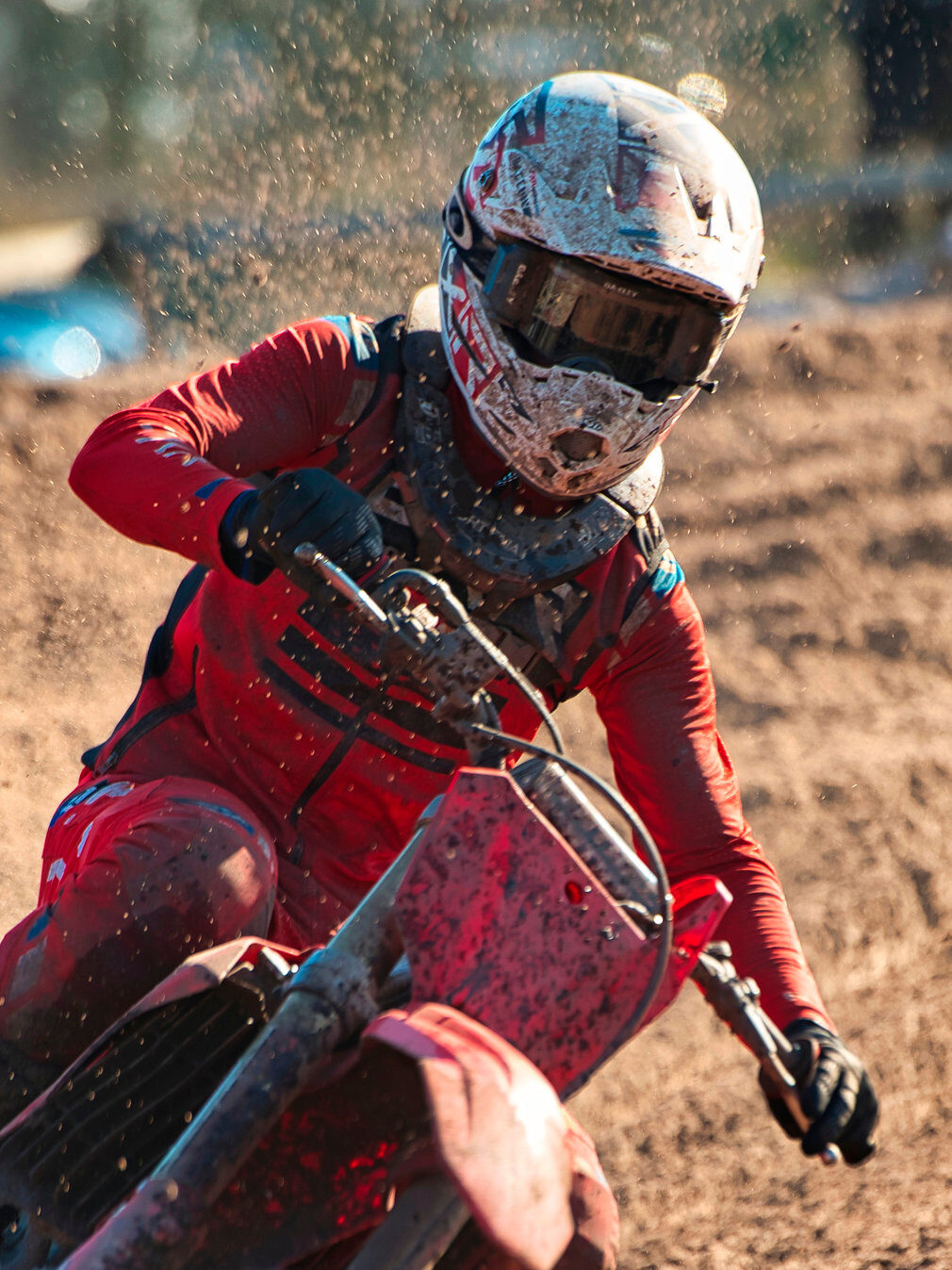 Ein Motocross-Fahrer in roter Montur und mit einem Helm, der mit Schlamm bedeckt ist, macht eine scharfe Kurve auf einer unbefestigten Strecke und fängt dabei herumfliegende Trümmer und intensive Action ein - perfekt, um Motocross-Sponsoring zu präsentieren. Rider's Point der Kaltenbach Gruppe. Dein Partner für BMW Motorrad und Honda.
