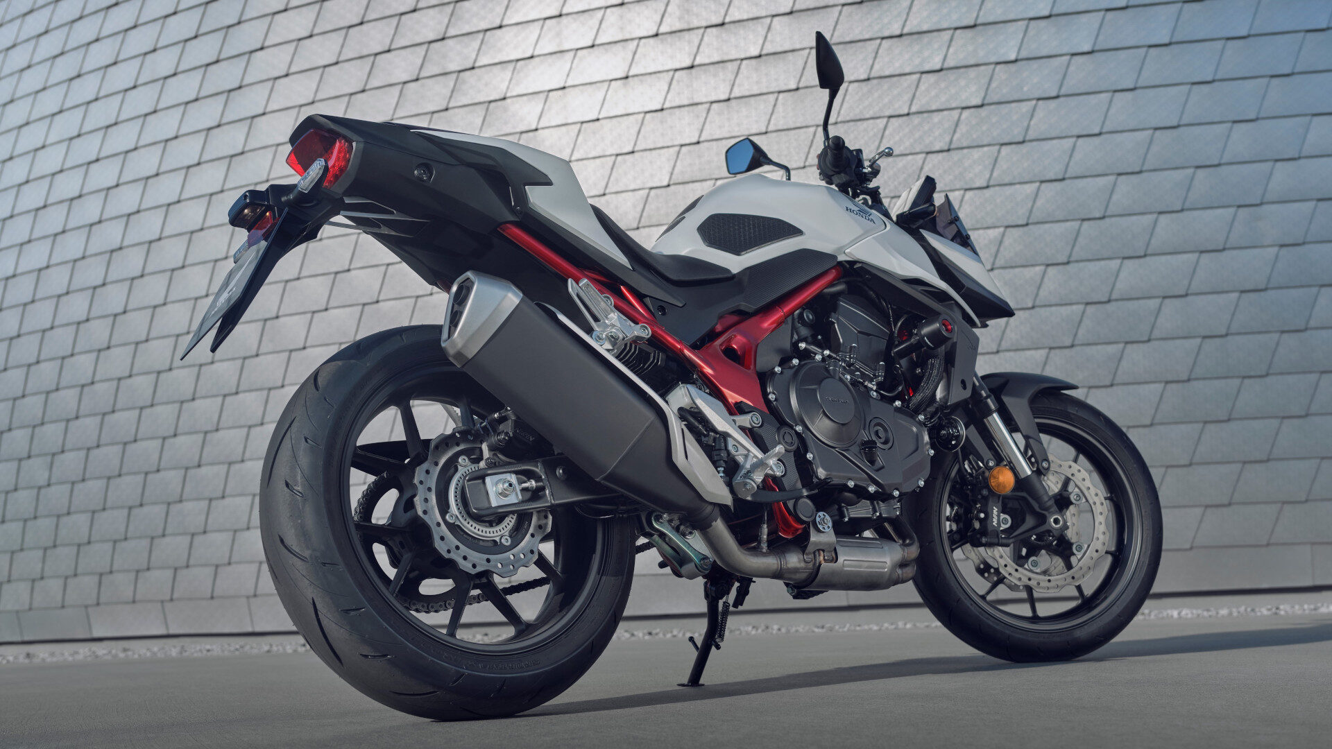 Ein modernes weiß-schwarzes Honda CB 750 Hornet Sportmotorrad mit roten Akzenten ist auf einer Betonfläche vor einer großen, metallischen Kachelwand geparkt. Der niedrige Aufnahmewinkel hebt das Heck und die Seite des Motorrads hervor. Rider's Point der Kaltenbach Gruppe. Dein Partner für BMW Motorrad und Honda.