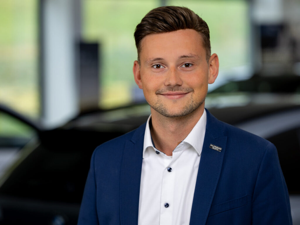 Ein junger Mann mit kurzen braunen Haaren, der eine blaue Anzugsjacke und ein weißes Hemd trägt, lächelt in die Kamera. Der Hintergrund ist unscharf und zeigt ein Auto in einem hellen, modernen Innenraum - perfekt für Ihren Honda Ansprechpartner. Rider's Point der Kaltenbach Gruppe. Dein Partner für BMW Motorrad und Honda.