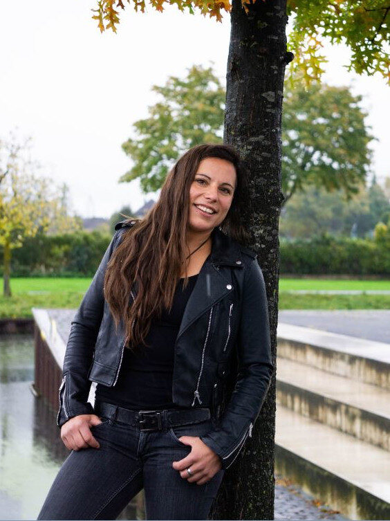 Eine Frau mit langen braunen Haaren, bekleidet mit einer schwarzen Lederjacke und schwarzen Jeans, lehnt an einem Baum und lächelt. Die Szene deutet auf ihren Kurven-Räubertochter-Geist hin - es gibt Grün, einen Spazierweg und Wasser im Hintergrund. Rider's Point der Kaltenbach Gruppe. Dein Partner für BMW Motorrad und Honda.