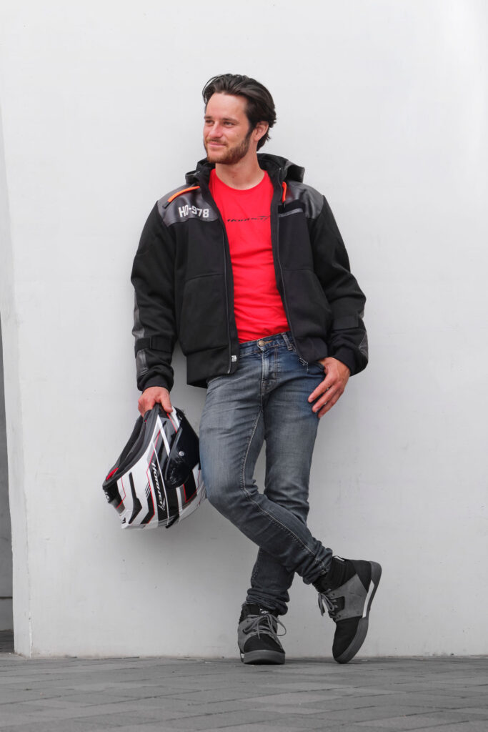 Ein Mann in schwarzer Jacke, rotem Hemd, Jeans und grauen Turnschuhen steht vor einer weißen Wand, hält einen Motorradhelm in der linken Hand und lächelt zur Seite - vielleicht denkt er über seinen Gutschein Motorradkauf nach. Rider's Point der Kaltenbach Gruppe. Dein Partner für BMW Motorrad und Honda.