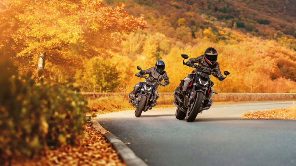 Zwei Motorradfahrer mit Helmen kurven am Rider's Point über die Straße, umgeben von leuchtendem Herbstlaub, goldenen und orangefarbenen Bäumen und einem bewaldeten Hügel im Hintergrund. Rider's Point der Kaltenbach Gruppe. Dein Partner für BMW Motorrad und Honda.
