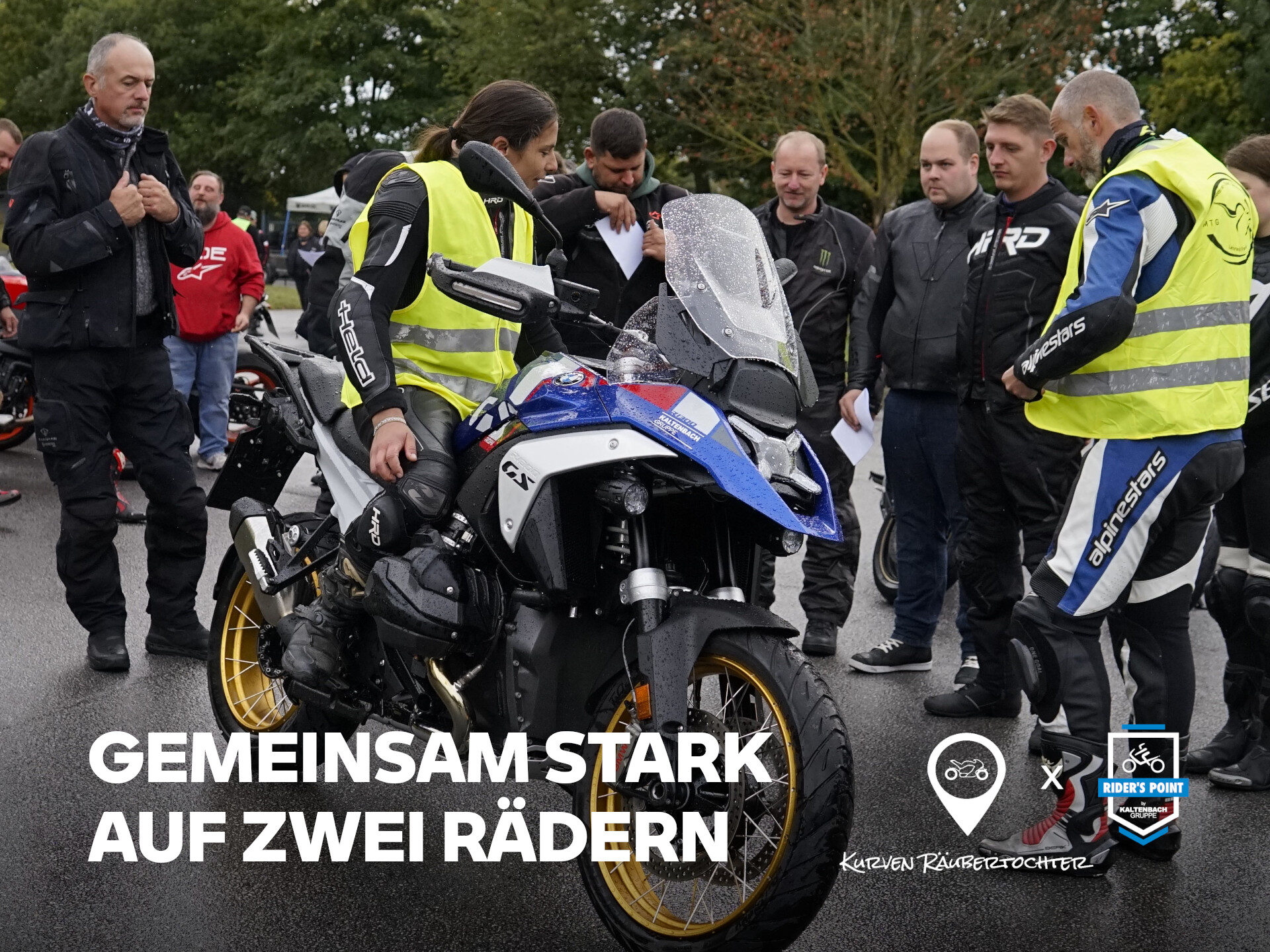 Eine Gruppe von Motorradfahrern, einige mit Warnwesten, versammelt sich um ein geparktes Motorrad auf einer nassen Straße - Motorradtraining in Aktion. Der Text lautet: GEMEINSAM STARK AUF ZWEI RÄDERN. Unten rechts sind zwei Logos zu sehen. Rider's Point der Kaltenbach Gruppe. Dein Partner für BMW Motorrad und Honda.