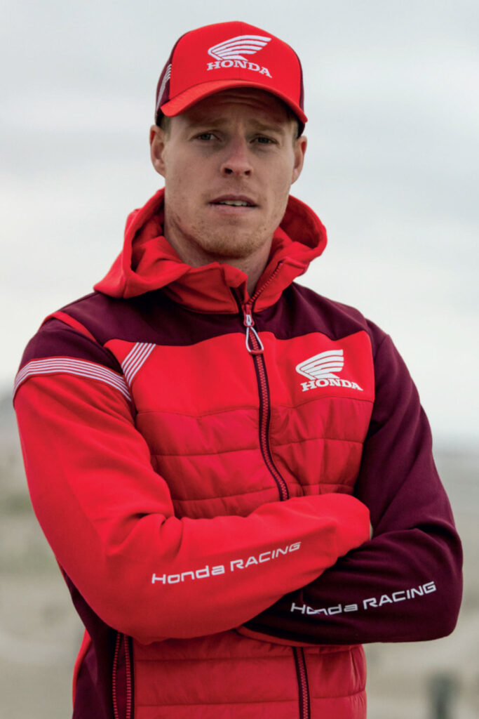Eine Person mit roter Honda Racing Jacke und passender roter Honda Mütze steht mit verschränkten Armen vor einem unscharfen Hintergrund im Freien und schaut in die Kamera - die perfekte Inspiration für Ihren nächsten Gutschein Motorradkauf. Rider's Point der Kaltenbach Gruppe. Dein Partner für BMW Motorrad und Honda.
