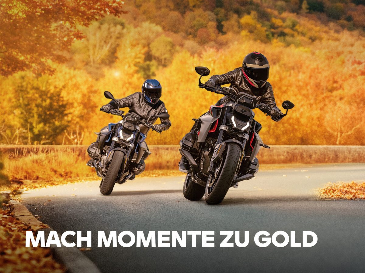 Zwei Motorradfahrer fahren auf einer kurvenreichen Straße, umgeben von leuchtenden Motorrad Herbst Farben und orangefarbenen Herbstblättern. Der Text "MACH MOMENTE ZU GOLD" wird am unteren Rand des Bildes angezeigt. Rider's Point der Kaltenbach Gruppe. Dein Partner für BMW Motorrad und Honda.